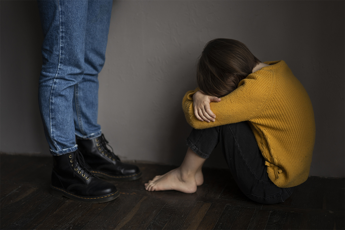 Pessoa sentada no chão, abraçada aos joelhos, ao lado de outra pessoa de pé
